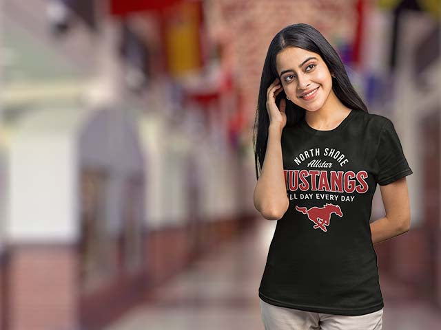 Woman wearing a black t-shirt with 'North Shore Mustangs' design, standing in a blurred indoor setting.
