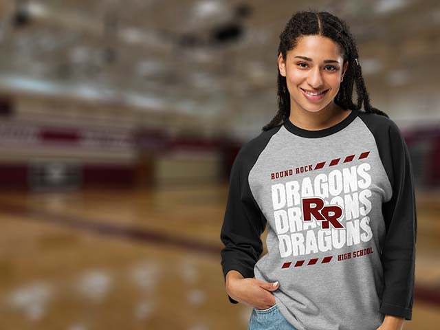 A smiling young woman stands in a gymnasium wearing a heather gray and black raglan baseball t-shirt with 'DRAGONS HIGH SCHOOL' and 'ROUND ROCK' printed on the front.