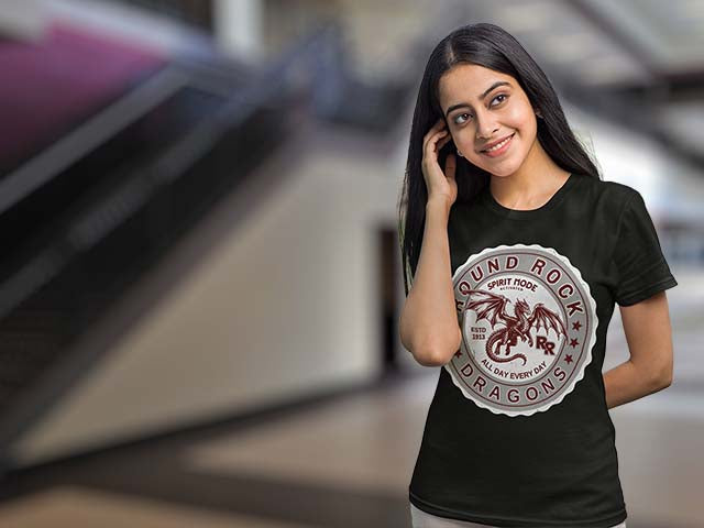 A woman wearing a black t-shirt with a circular graphic that reads 'ROUND ROCK SPIRIT ROCK DRAGONS' around a red dragon illustration, set against a blurred indoor background.