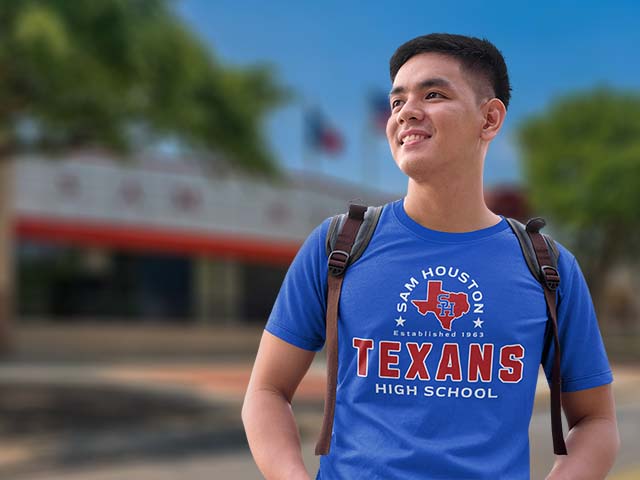 A smiling young man wearing a blue Sam Houston High School Texans t-shirt and a backpack, standing outdoors with a blurred school building and trees in the background.