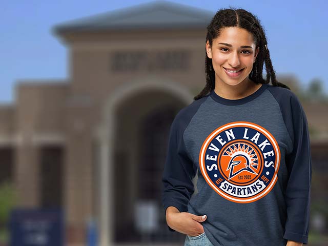 A smiling young woman stands outdoors in front of a blurred building, wearing a navy and slate blue raglan long-sleeve shirt with a large circular 'Seven Lakes Spartans' logo on the chest.