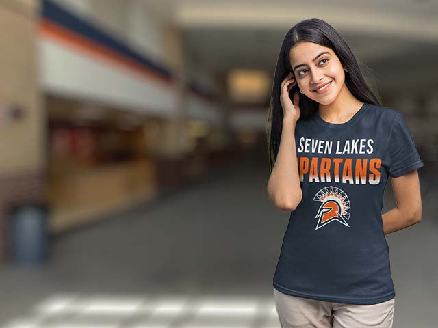 A woman wearing a navy blue t-shirt with 'SEVEN LAKES SPARTANS' and a Spartan logo printed on the front, standing in a blurred indoor hallway.