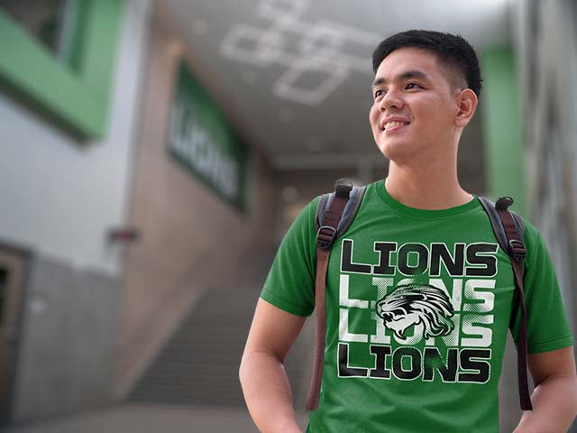 A smiling young man wearing a green t-shirt with a 'LIONS' graphic, standing in a blurred outdoor corridor. Spring High School Lions Classic T-shirts All Products Page