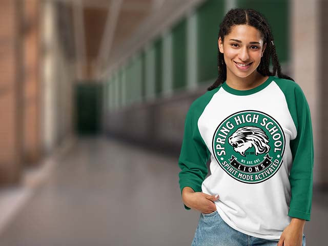 A smiling young woman stands in a blurred indoor hallway wearing a green and white raglan baseball-style shirt with a large circular 'Spring High School' graphic on the front, paired with blue jeans.