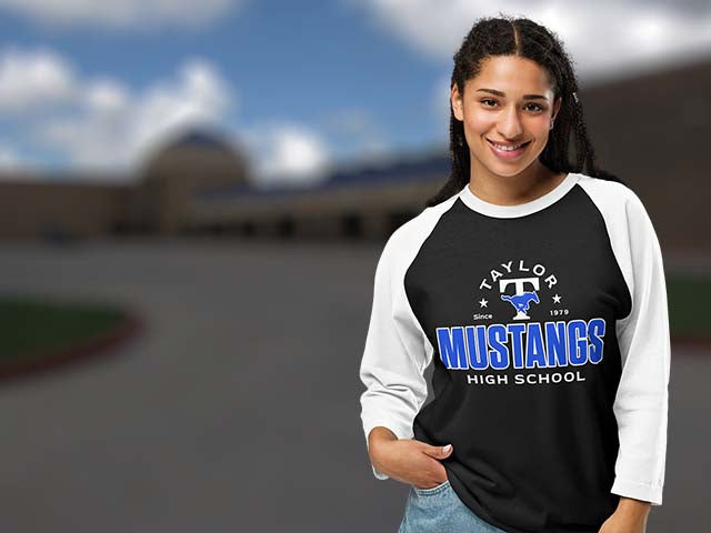 A smiling young woman wearing a black and white raglan baseball t-shirt with 'Taylor Mustangs High School' printed in blue and white on the front, set against a blurred outdoor school background.