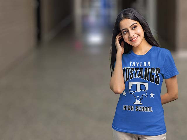 A woman wearing a bright blue short-sleeve T-shirt with 'TAYLOR MUSTANGS HIGH SCHOOL' and a large 'T' graphic printed on the front, standing in a blurred indoor hallway.