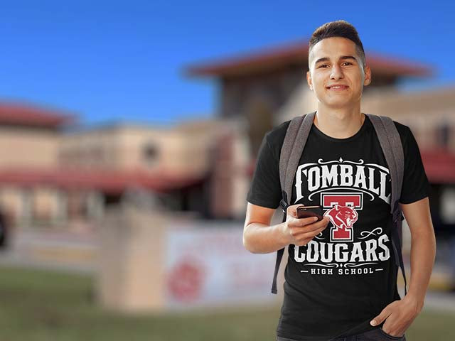 A young man wearing a black 'Tomball Cougars High School' graphic t-shirt with a gray backpack, holding a smartphone in front of a blurred outdoor school background.