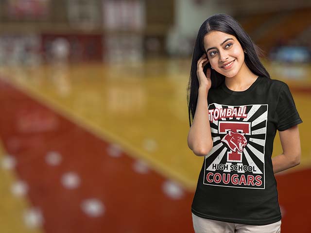 A woman standing on a blurred indoor basketball court wearing a black graphic t-shirt with 'Tomball High School Cougars' and a large cougar logo printed on the front. Tomball High School Cougars Women's T-shirts All Products Page