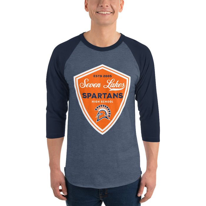 A man wearing a 3/4 sleeve raglan t-shirt with a heather slate blue body and navy blue sleeves. The shirt features a large orange, white, and navy shield graphic on the chest that reads 'Seven Lakes Spartans High School' with a Spartan helmet logo. Seven Lakes High School Spartans Unisex 3/4 sleeve Raglan T-shirt 225